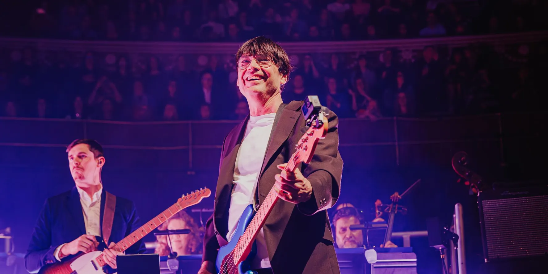 Alex James performing on bass with a full orchestra under purple stage lighting on the Britpop Classical tour.