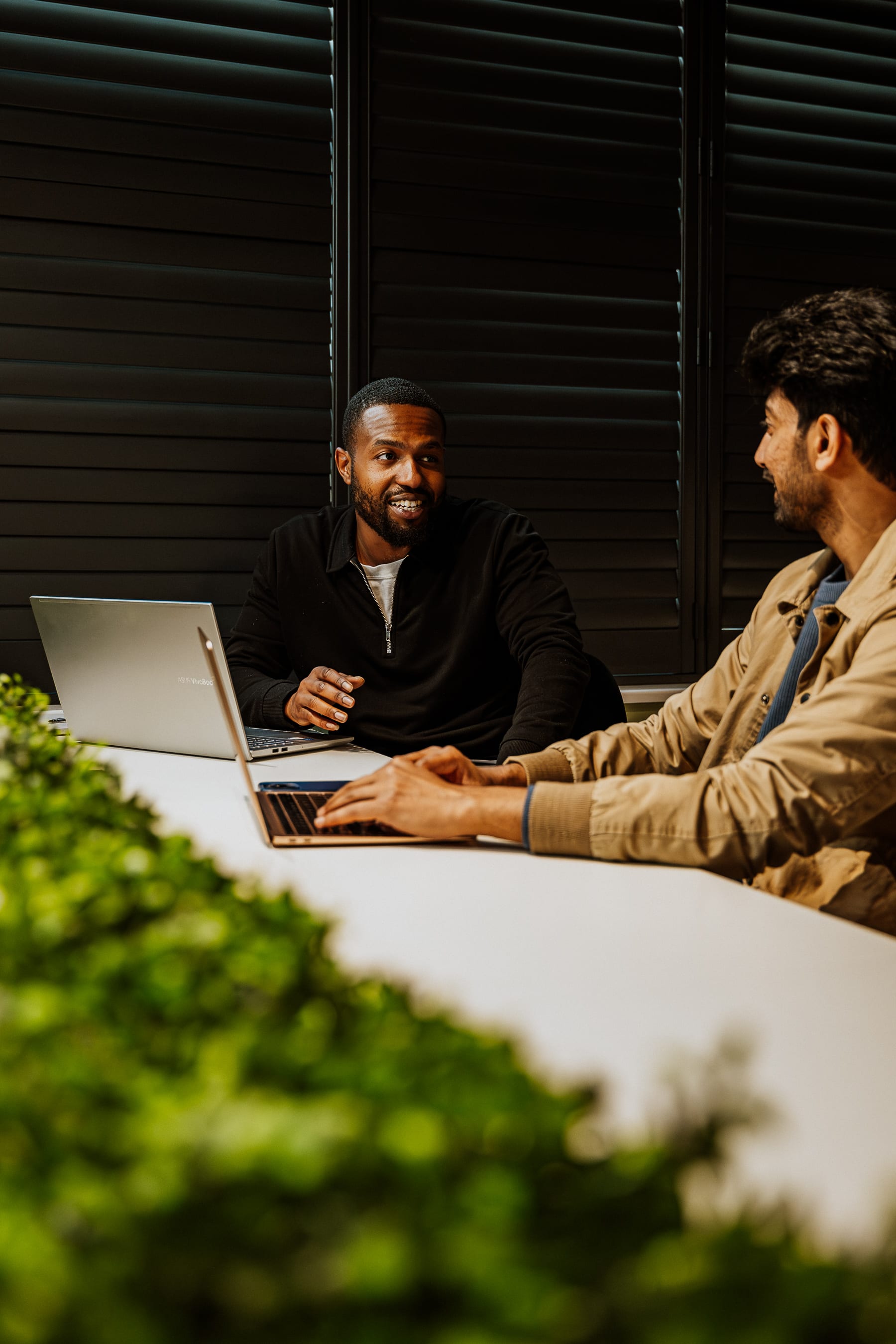 Two 21 Degrees team members talking over laptops at a meeting table, with office greenery in the foreground.