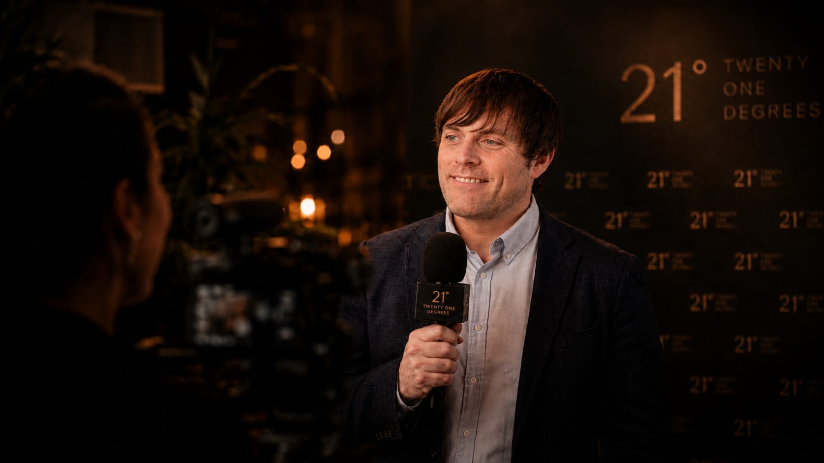 Jon Corbett being interviewed at a 21 Degrees event, holding a branded microphone and smiling in front of a dark 21 Degrees backdrop under warm lighting.